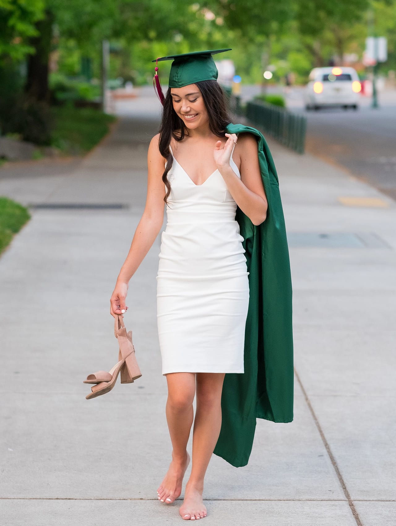 Mujer con vestido de graduación blanco satinado, birrete verde y toga al hombro durante una ceremonia de graduación al aire libre.