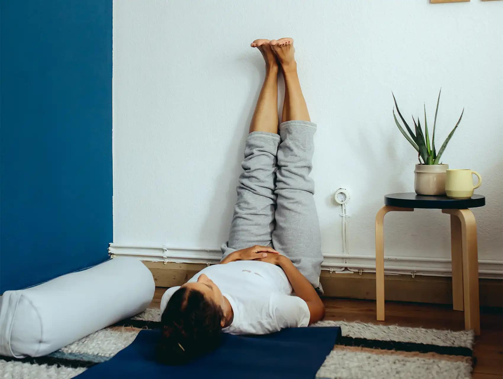 Woman practicing vertical leg extension with white bolster beside her yoga mat with bolsters