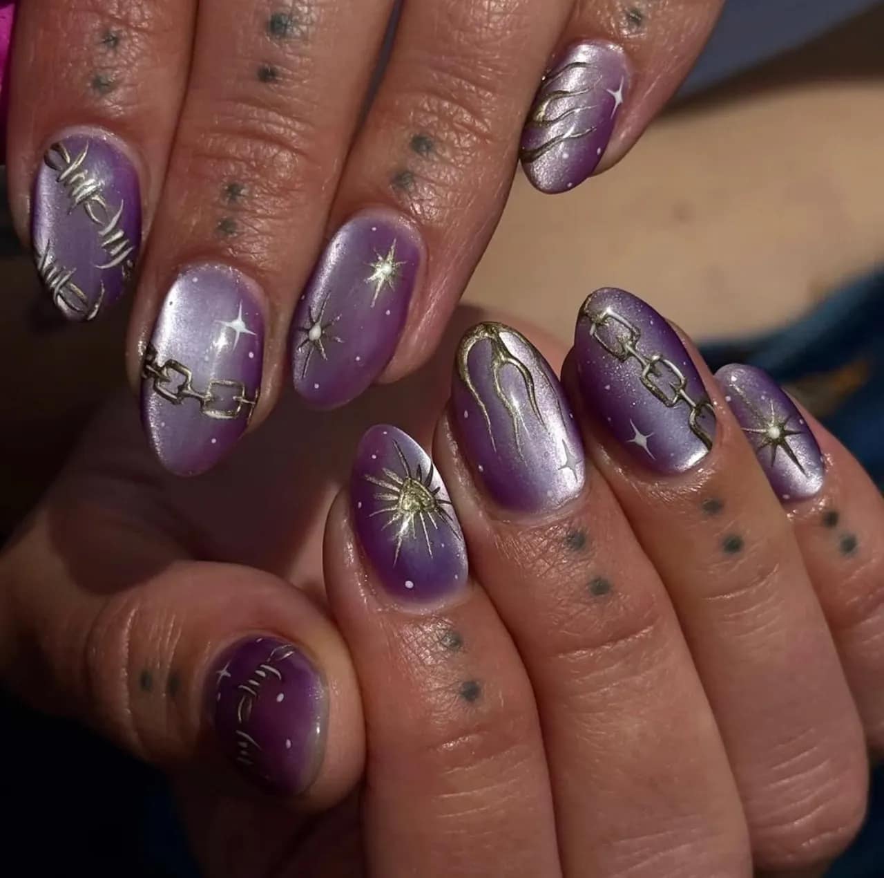 Diseños de uñas acrílicas moradas con detalles dorados de fantasía como cadenas, estrellas y símbolos místicos.