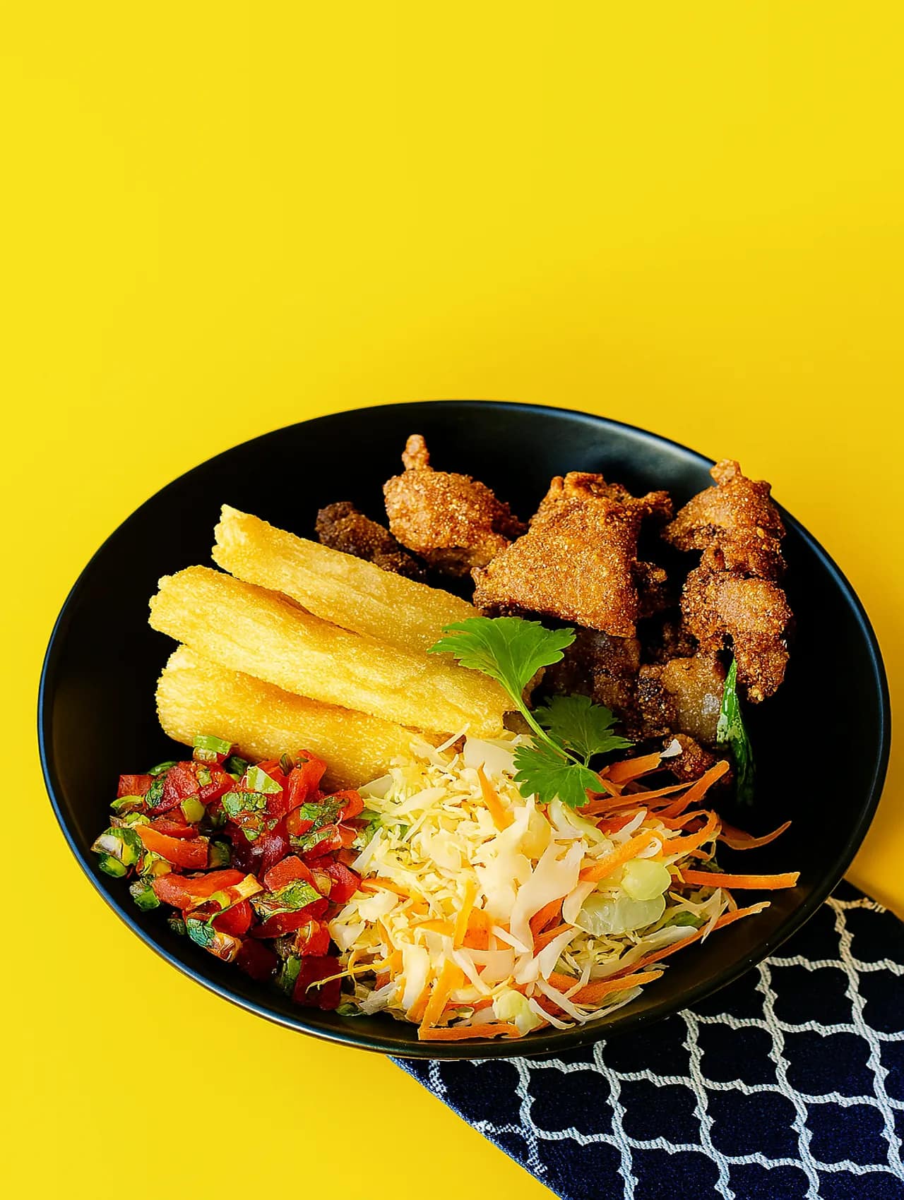 Plato con chicharrones crujientes, yuca frita, curtido de repollo y pico de gallo servido en plato negro sobre fondo amarillo.