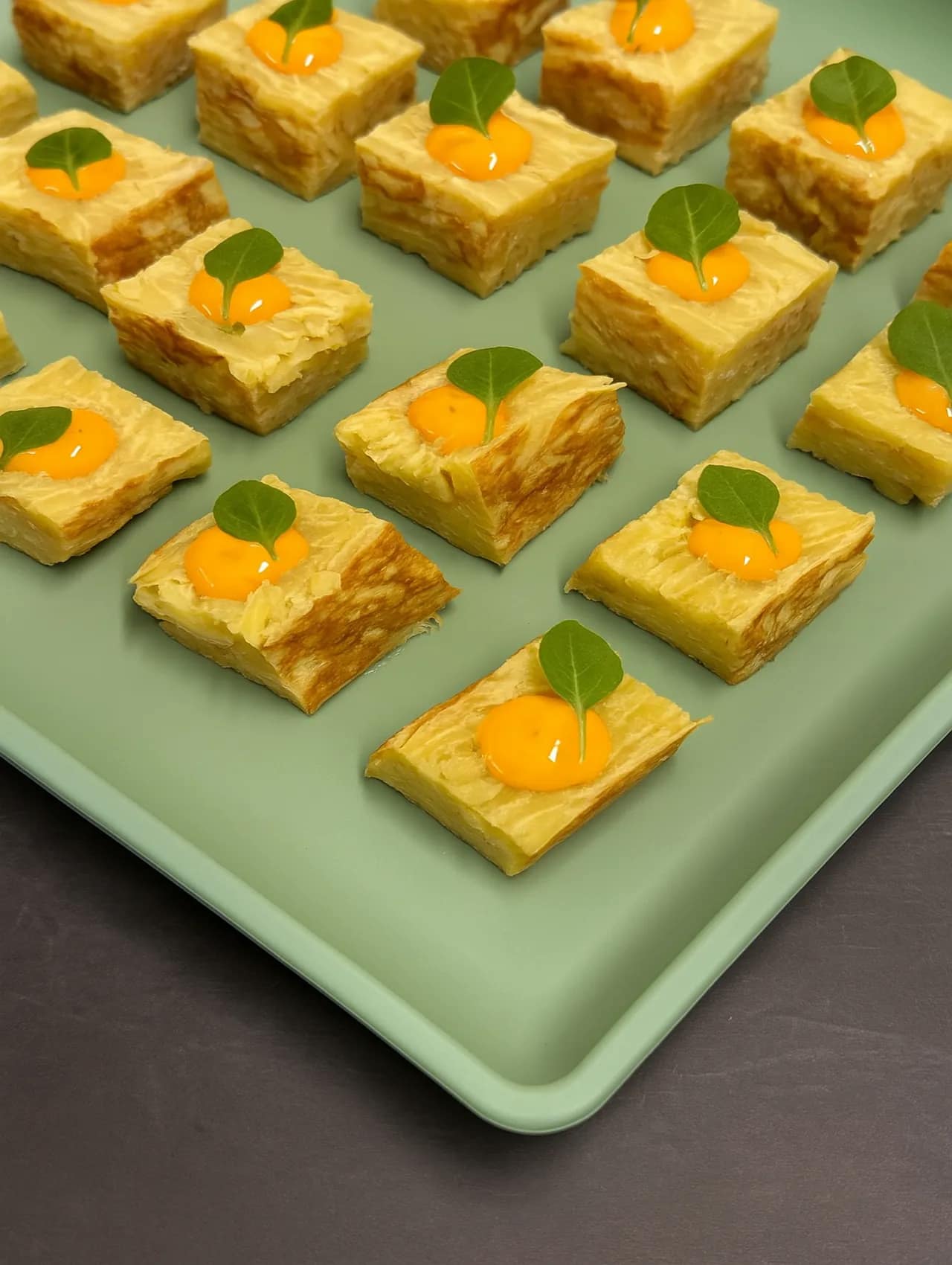 Cubitos de tortilla española con un toque de alioli y hoja verde sobre bandeja verde.